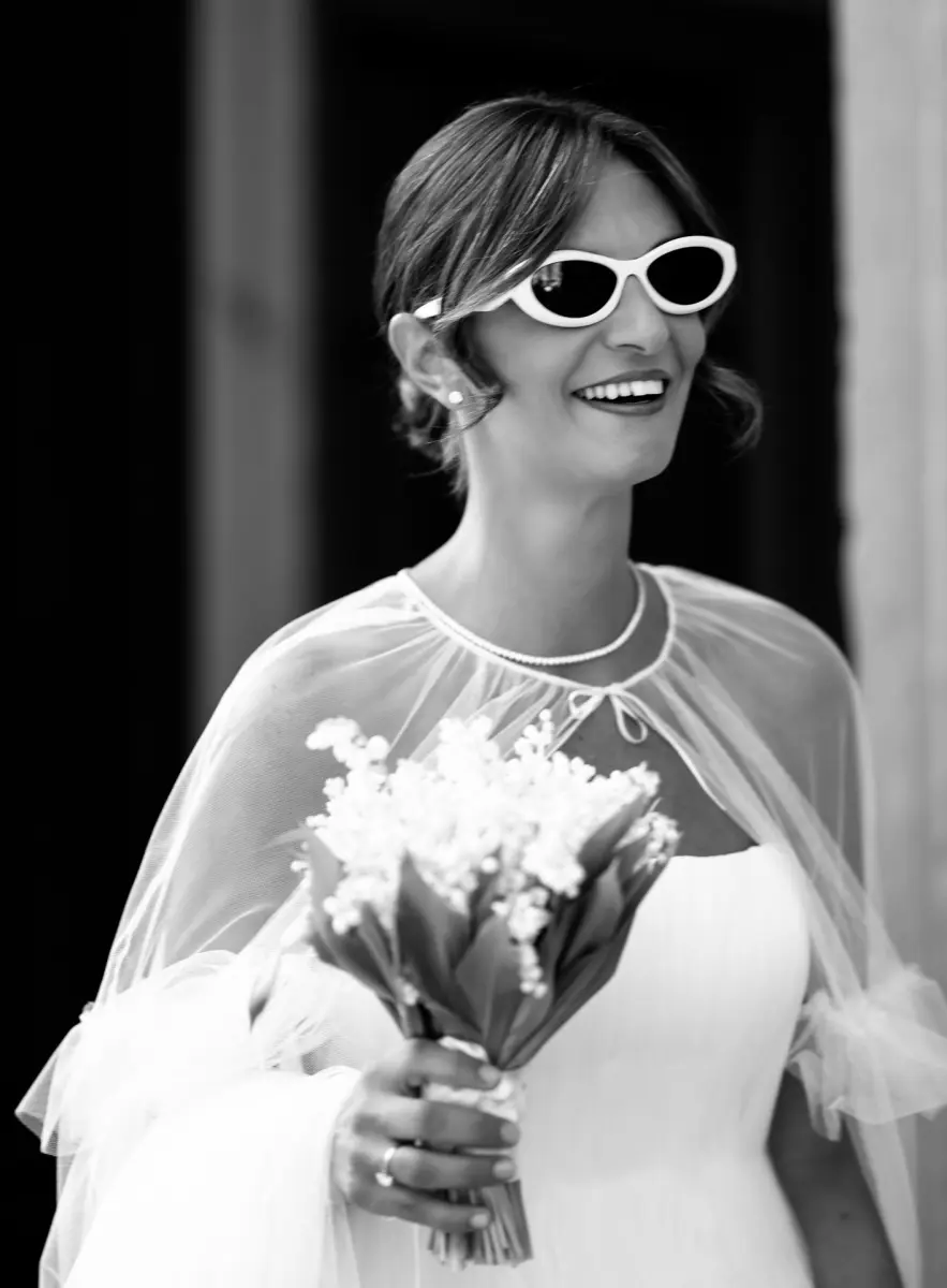Una sposa ride con gli occhiali da sole bianchi e il mughetto in mano, in bianco e nero
